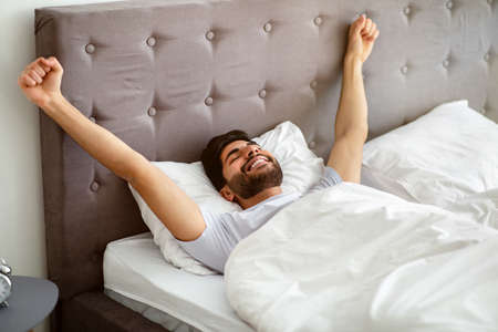 Wellslept arab guy waking up and stretching hands, lying in comfortable bed with eyes closed