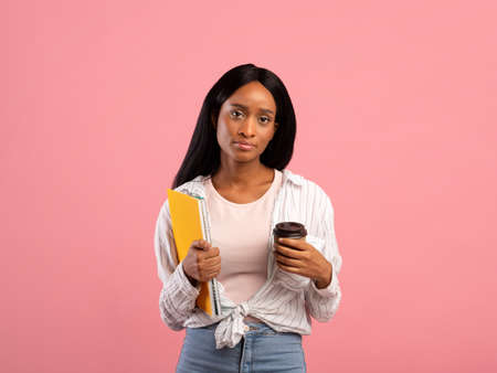 Tired black female student with notebooks and takeaway coffee looking at camera on pink studio backgroundの写真素材