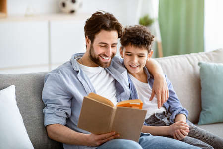 Leisure time. Happy father and son reading book and smiling, resting together on comfy sofaの写真素材