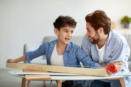 Best team. Cheerful father and son measuring plank of wood with tape. Family making birdhouse from wooden detailsの写真素材