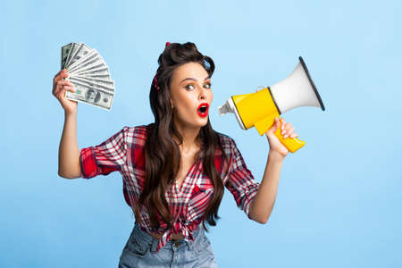 Pretty pin up woman holding fan of money and shouting into megaphone on blue studio backgroundの写真素材