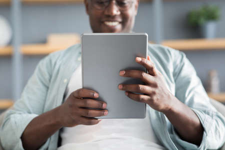 Free time concept. Black man using digital tablet, reading news or browsing internet, resting on the...