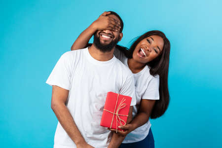 Celebration Concept. Excited African American woman covering her boyfriend's eyes, holding wrapped gift box and greeting him with birthday or anniversary, standing isolated over blue studio backgroundの写真素材