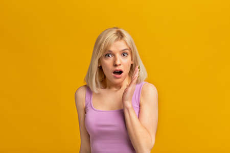 Dont tell anybody. Shocked blonde woman keeping hand near opened mouth telling secret over yellow studio backgroundの写真素材