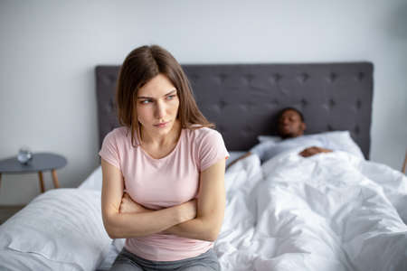 Depressed young woman having relationship problem, sitting on bed while her husband sleeping at...