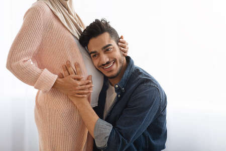 Happy eastern man embracing tummy of his pregnant wife, listening baby kicksの写真素材
