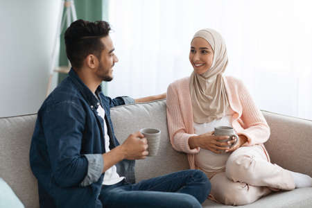 Cheerful Pregnant Muslim Couple Resting Together At Home, Drinking Coffee And Chattingの写真素材