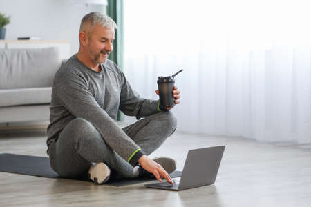 Senior man in sportswear sitting on fitness mat, using laptopの写真素材