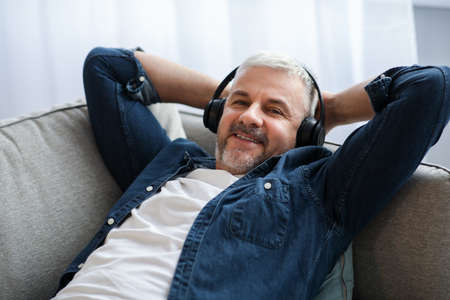 Happy matured man reclining on couch, listening to musicの写真素材