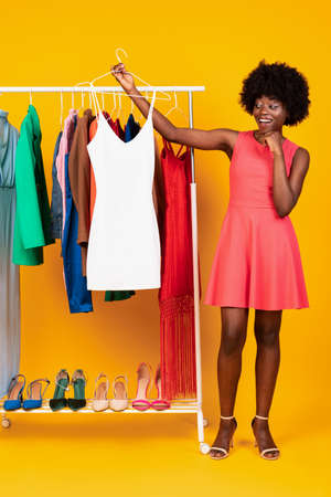 Black Woman Choosing Dress Standing Near Clothing Rail, Yellow Background