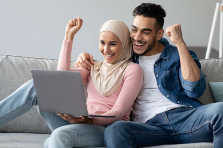 Emotional arab couple with laptop raising fists up, celebrating successの写真素材
