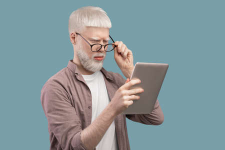 Strange content. Surprised albino man in glasses looking in digital tablet and reading news on turquoise backgroundの写真素材