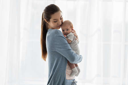 Beautiful young mother holding her adorable newborn baby while standing near window