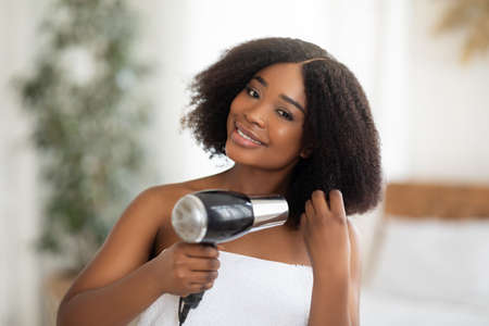 Beautiful black woman in bath towel using hairdryer after shower at homeの写真素材