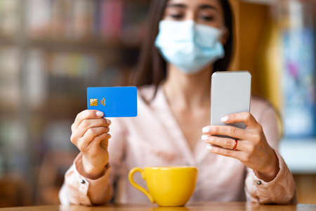 Contactless payment during virus quarantine. Woman in protective mask holding cellphone and credit...