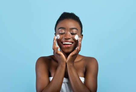 Joyful black woman applying hydration cream on cheeks, standing with closed eyes over blue studio...