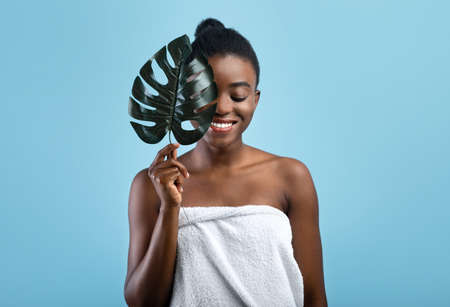 Pretty black woman with smooth skin wrapped in towel hiding behind tropical plant on blue studio backgroundの写真素材