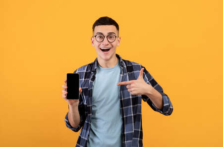 Excited millennial guy in casual wear pointing at smartphone with empty screen on orange background,...