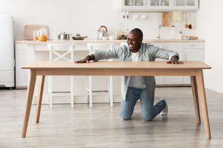 Overjoyed black man touching new wooden table, after assembing furniture with own hands at home in kitchen interiorの写真素材