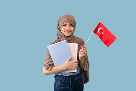 Teen girl in traditional Muslim hijab holding Turkish flag and textbooks on blue background. Education for Arab femalesの写真素材