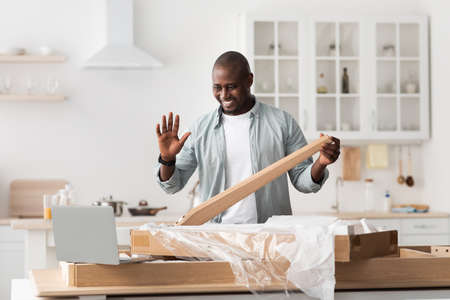 Black handyman shooting video instructions for assembling furniture at home with own hands, waving to laptop at homeの写真素材