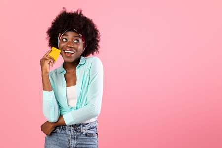 Cheerful Black Lady Holding Credit Card Looking Aside, Pink Background