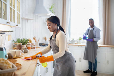 Cheerful black woman cleaning kitchen table, her boyfriend washing dishes on background, blank space