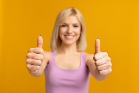 Best choice. Smiling woman showing thumbs up gesture with both hands, posing over yellow studio...