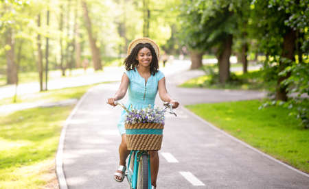 Joyful African American lady in blue dress bicycling at park in summertimeの写真素材