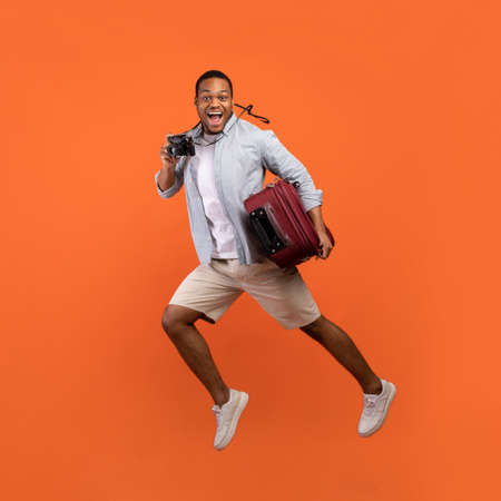 Full length portrait of excited black guy with suitcase and camera jumping on orange studio...