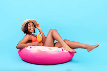 Sexy black lady sitting on inflatable donut ring, drinking summer cocktail on blue background, full...