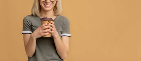 Favorite hot drink. Unrecognizable happy lady holding takeaway paper cup, isolated on beige background with free spaceの写真素材