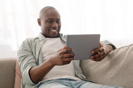 Positive african american mature man spending time with digital tablet, browsing internet while resting on comfy couchの写真素材