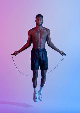 Full length portrait of handsome black sportsman with naked torso jumping on skipping rope in neon lightingの写真素材