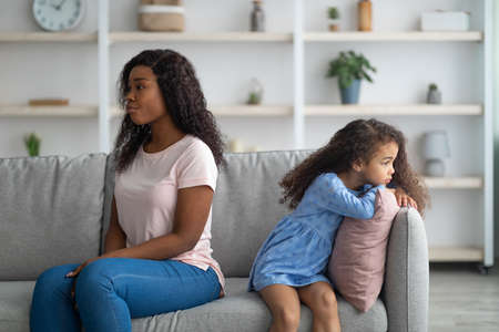 Offended black woman and her daughter sitting back to back on sofa, not speaking to each other after fight at homeの写真素材
