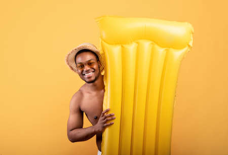 Young black guy wearing straw hat and sunglasses, holding inflatable lilo on yellow studio background. Summer vacationの写真素材