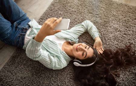 Happy Indian lady in headphones lying on floor and listening to music on mobile device at home, above viewの写真素材