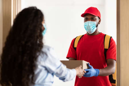 Black delivery man in medical mask giving box to woman