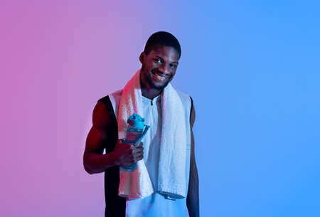 Young black sportsman with bath towel holding bottle of water in neon lightの写真素材