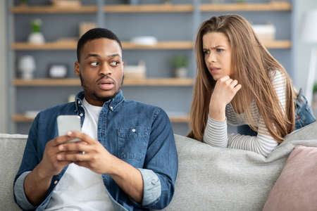 Infidelity Concept. Suspicious Young Woman Watching Her Black Boyfriend Texting On Phone