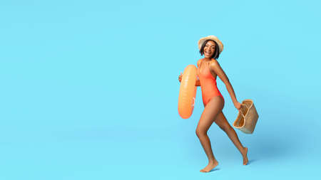 Hello summer. Positive black lady in bikini doing to beach with straw bag and inflatable ring on blue studio backgroundの写真素材