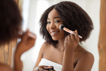 Natural makeup for young skin. Sexy black lady putting on blusher or powder with brush in front of mirror indoorsの写真素材