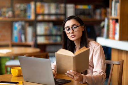 Thoughtful latin woman reading book in front of laptop at cafeteria, studying and getting ready for...
