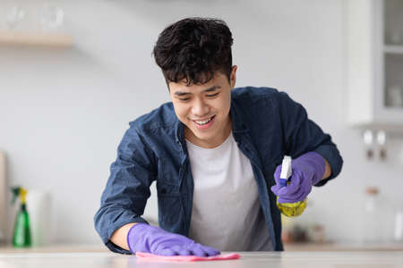 Cheerful asian guy cleaning kitchen table with spray and clothの写真素材