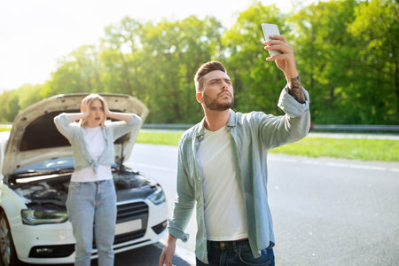 Sad young man standing on road next to broken car together with girlfriend, trying to call...
