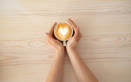 Woman holding cup of coffee latte, with heart shape, enjoying hot beverage in cafeteria, top view