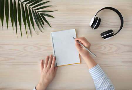 To do list. Female hands writing with pencil in notebook , sitting on wooden table with headphones...