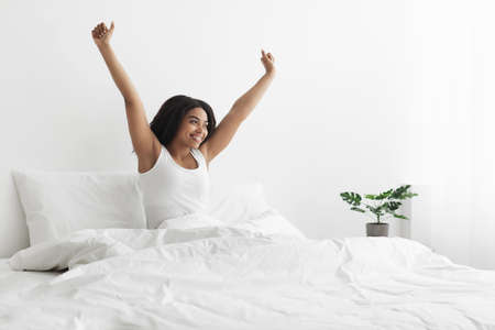 Relaxed after good night sleep. Happy african american woman sitting on bed with blanket, stretching...