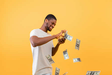 Wealth concept. Young rich black guy throwing out money bills, standing over yellow studio...