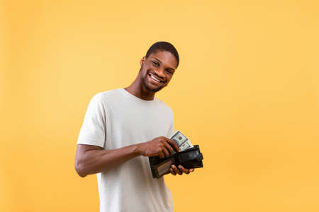 Investment and money saving concept. Happy african american guy putting banknotes into wallet over yellow backgroundの写真素材